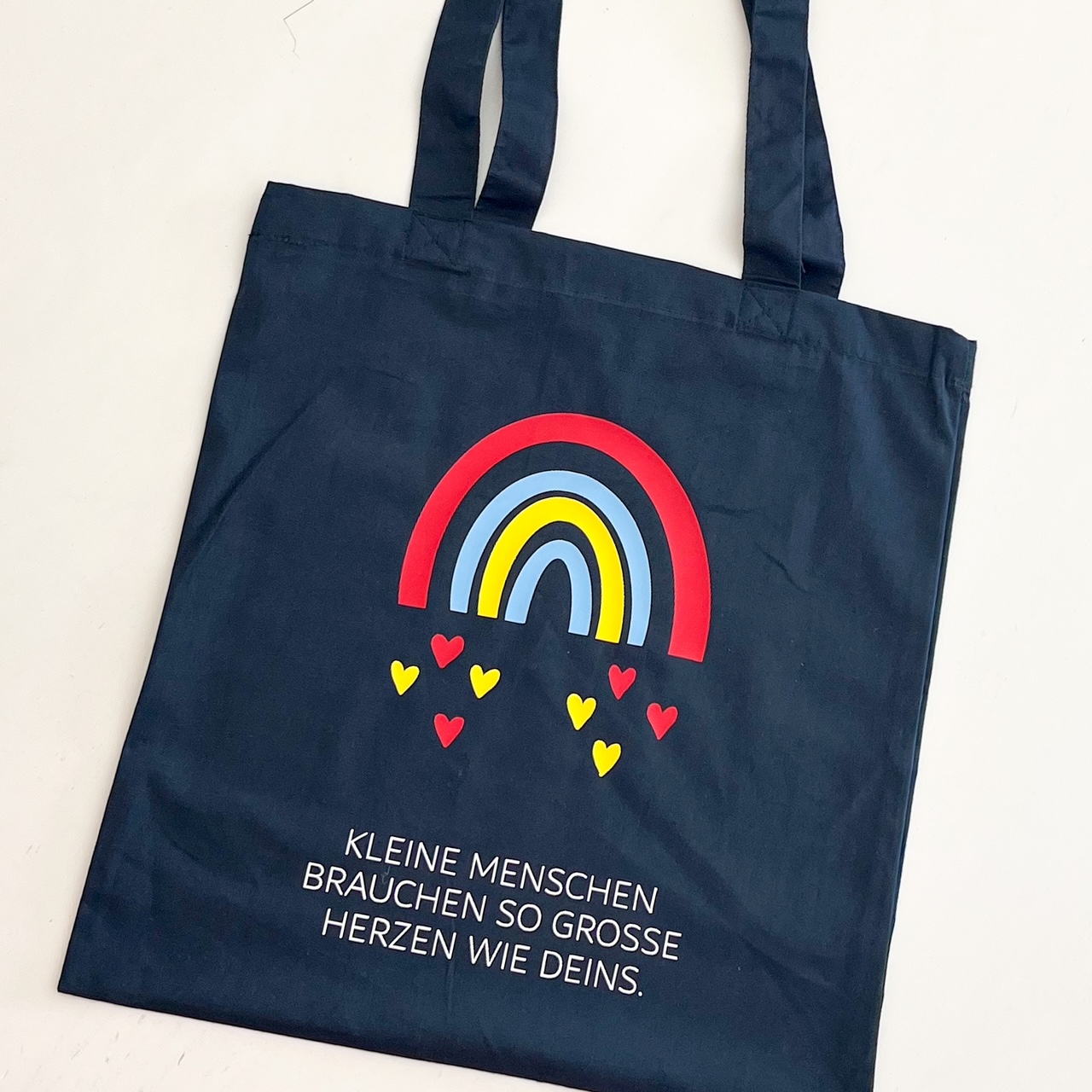 Dunkelblaue Stofftasche mit langem Henkel und einem Regenbogen in den Farben rot, hellblau, gelb. Der Spruch unter dem Regenbogen in weiß. Der Spruch: Kleine Menschen Brauchen so grosse Herzen wie deins.